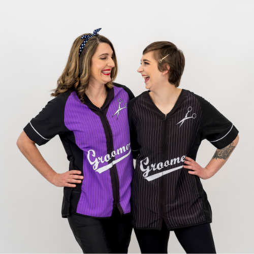 Two groomers smiling at each other, the one on the left wearing a Purple Baseball smock, and the one on the left wearing a Black Baseball smock.