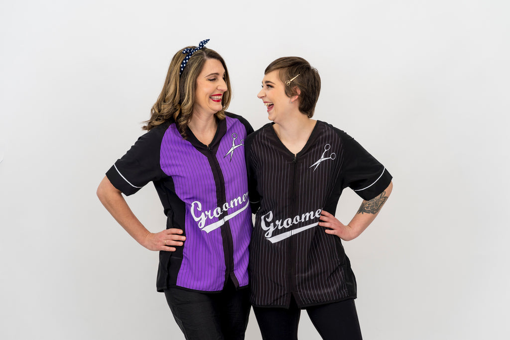 2 dog groomers smiling at each other while wearing grooming smocks , one on the left wearing a Purple Baseball grooming smock, One on the right wearing a Black Baseball grooming smock